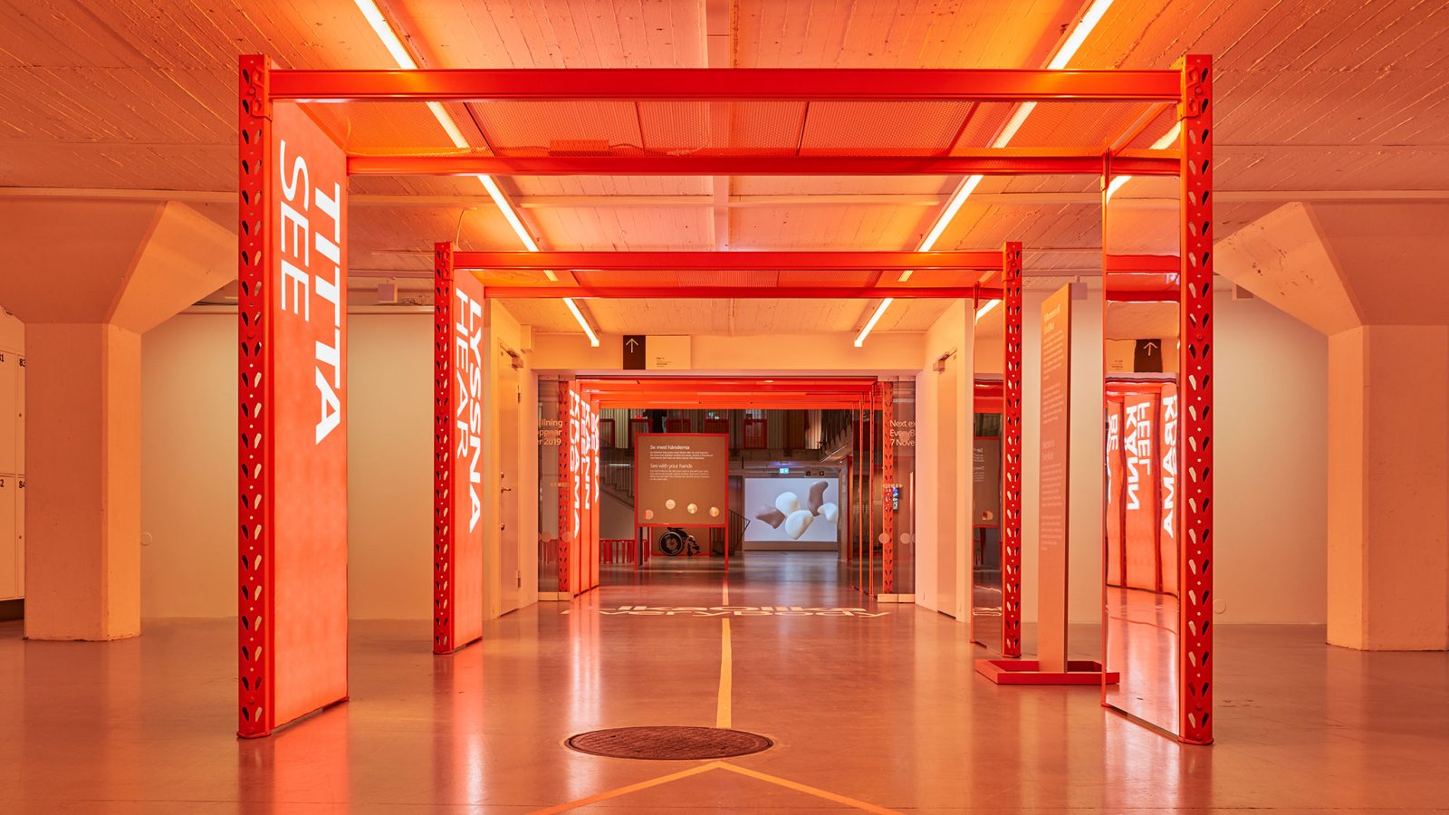 En korridor inne på IKEA Museum med orange belysning och flera orange pelare med stora, vita bokstäver som bildar orden ”TITTA”, ”LYSSNA” o.s.v.