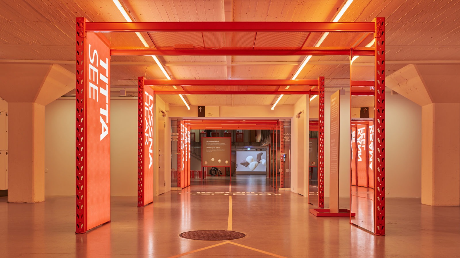 A corridor inside IKEA Museum with orange light and several orange arches with large, white writing saying ‘SEE’, ‘HEAR’ etc.