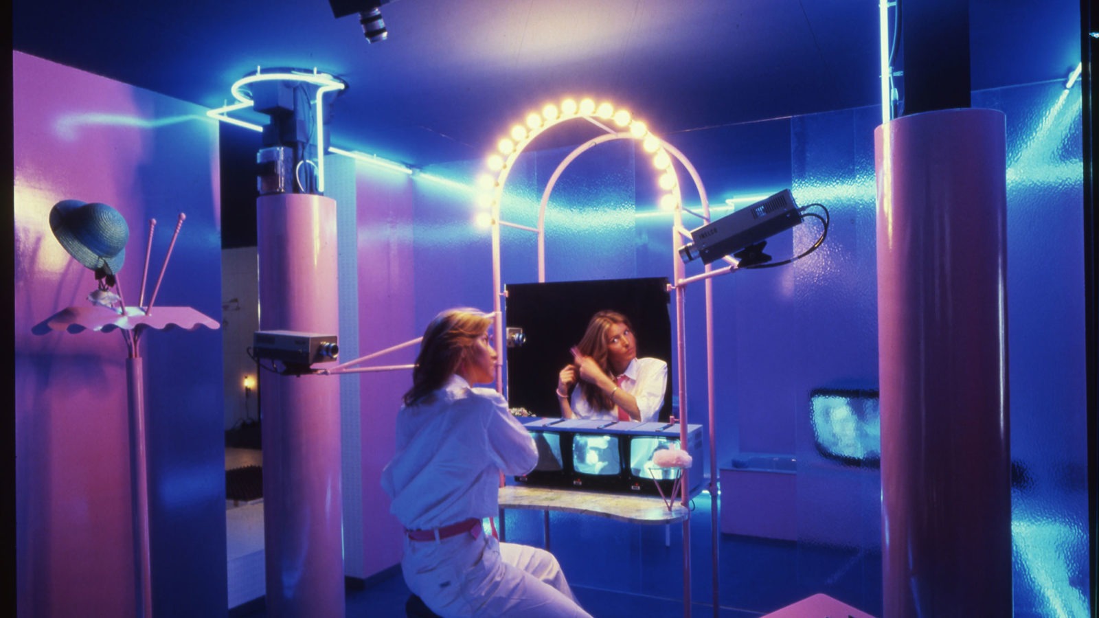 A woman combing her hair and looking at a mirror in a lilac-blue room dimly lit with neon lights and cameras pointed at her.