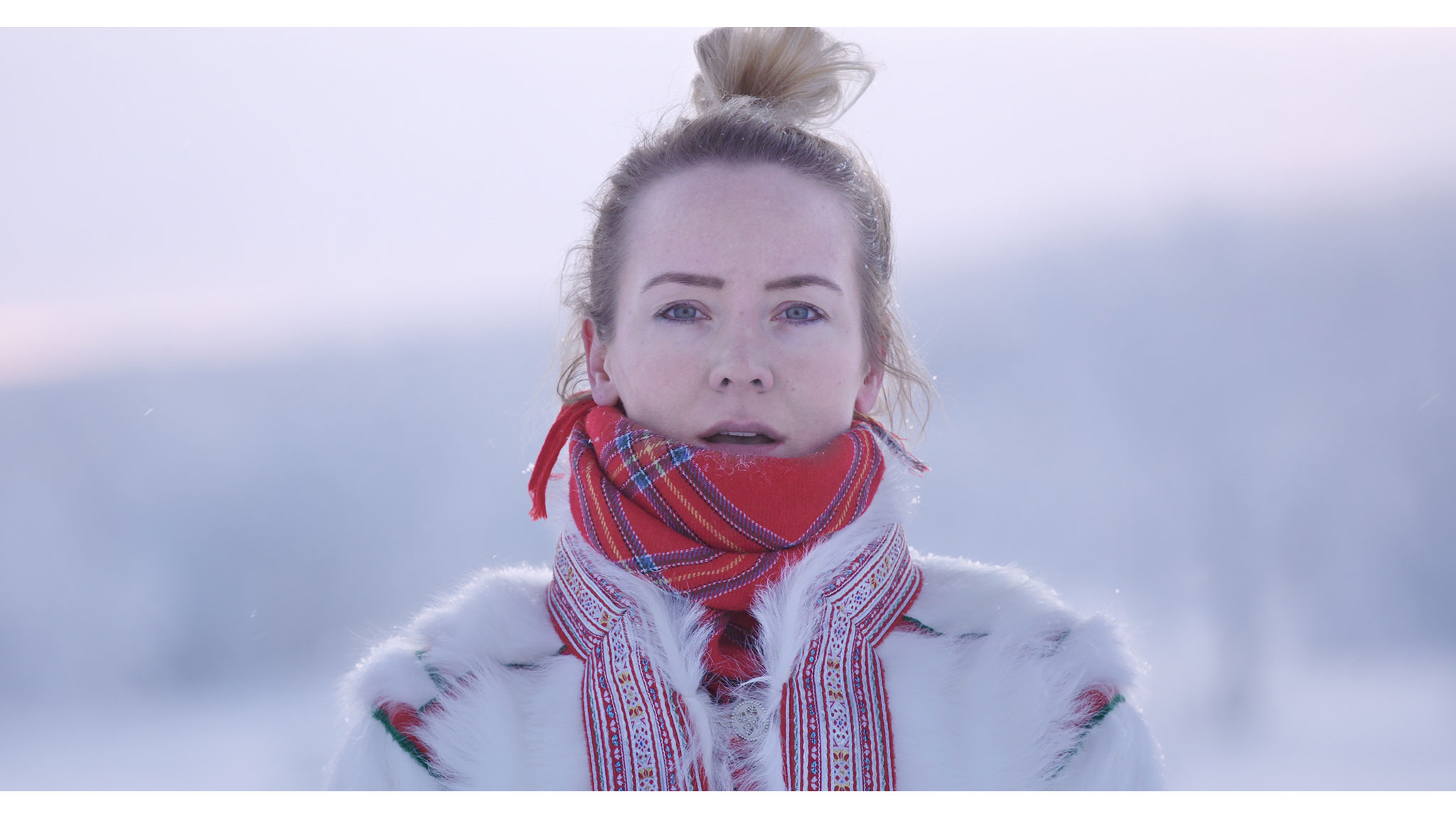 En ung kvinna i ett vinterlandskap klädd i traditionell samisk jacka av renskinn och en röd scarf.