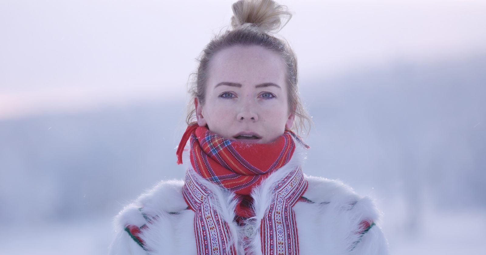 A young woman in a snowy winter landscape wearing a traditional Lapp reindeer jacket and a red scarf.