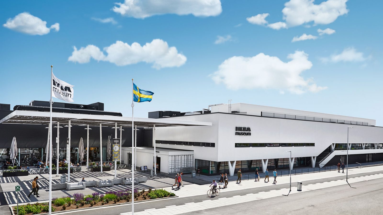 Den vita byggnaden som inrymmer IKEA Museum, under en blå himmel, människor går omkring utanför och en IKEA flagga och en svensk flagga vajar i vinden utanför byggnaden.