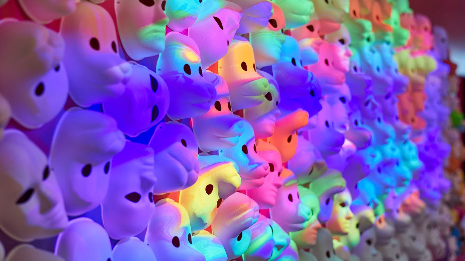 Face masks for play covering a large wall completely, bathed in multicoloured, fluid light.