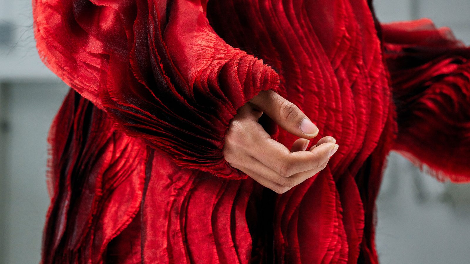 Ein rotes Kleidungsstück mit langen Ärmeln und mehreren senkrecht fallenden Lagen mit gewellter Textur, Schatten und eine Frauenhand.