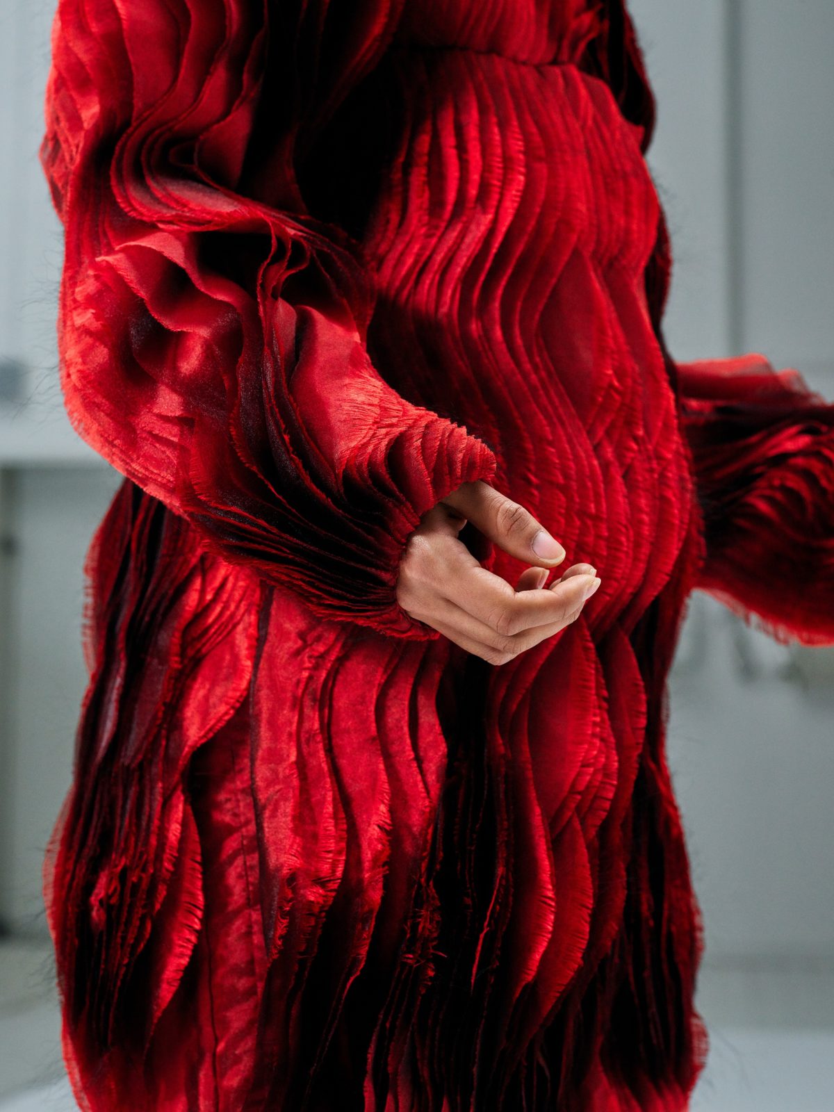 A red piece of clothing with long sleeves and multiple vertical layers with a wavy texture, shadows, and a woman’s hand.