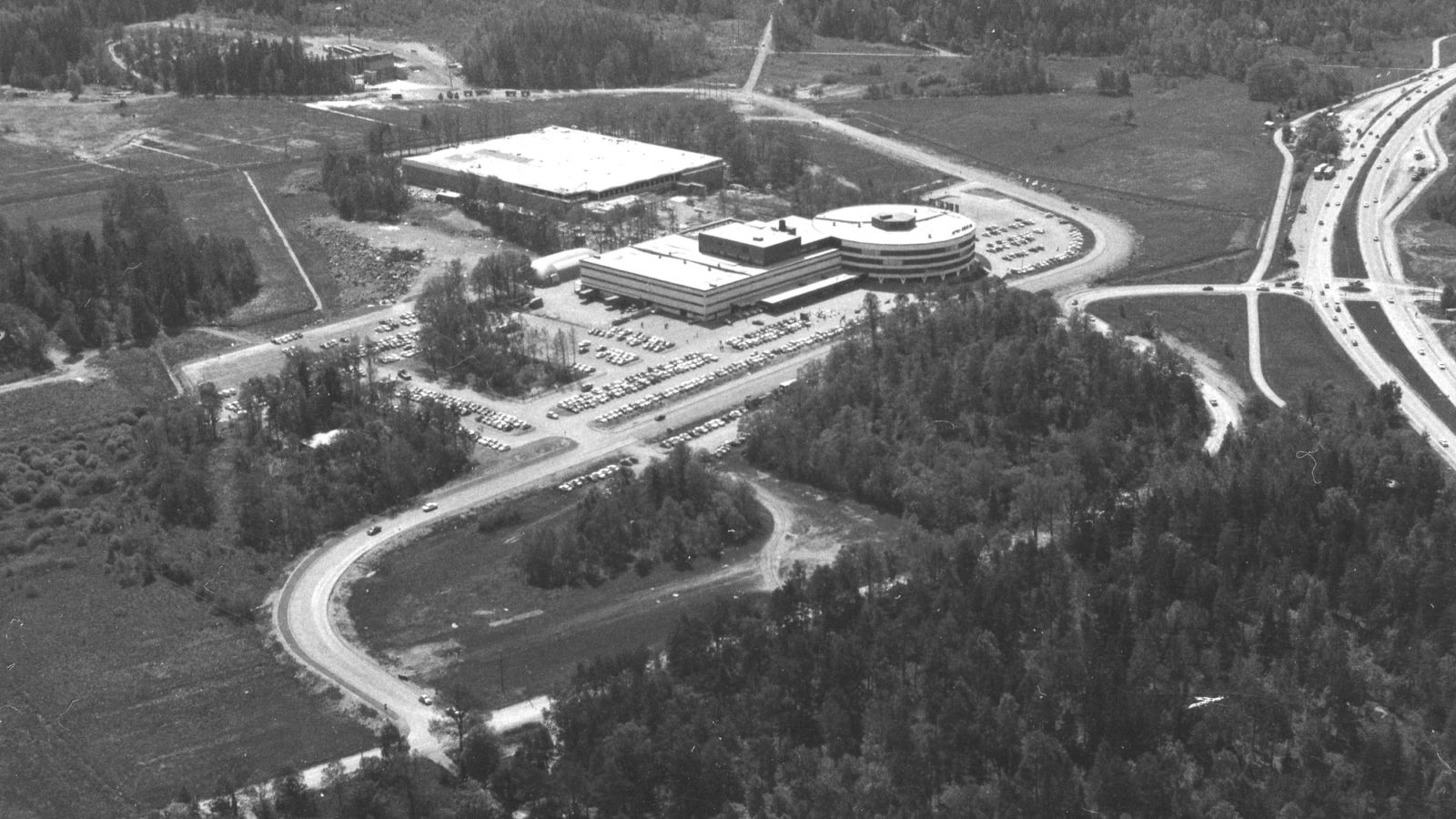 Black and white aerial photo of huge buildings surrounded by parking lots, roads, forests, fields and highways.