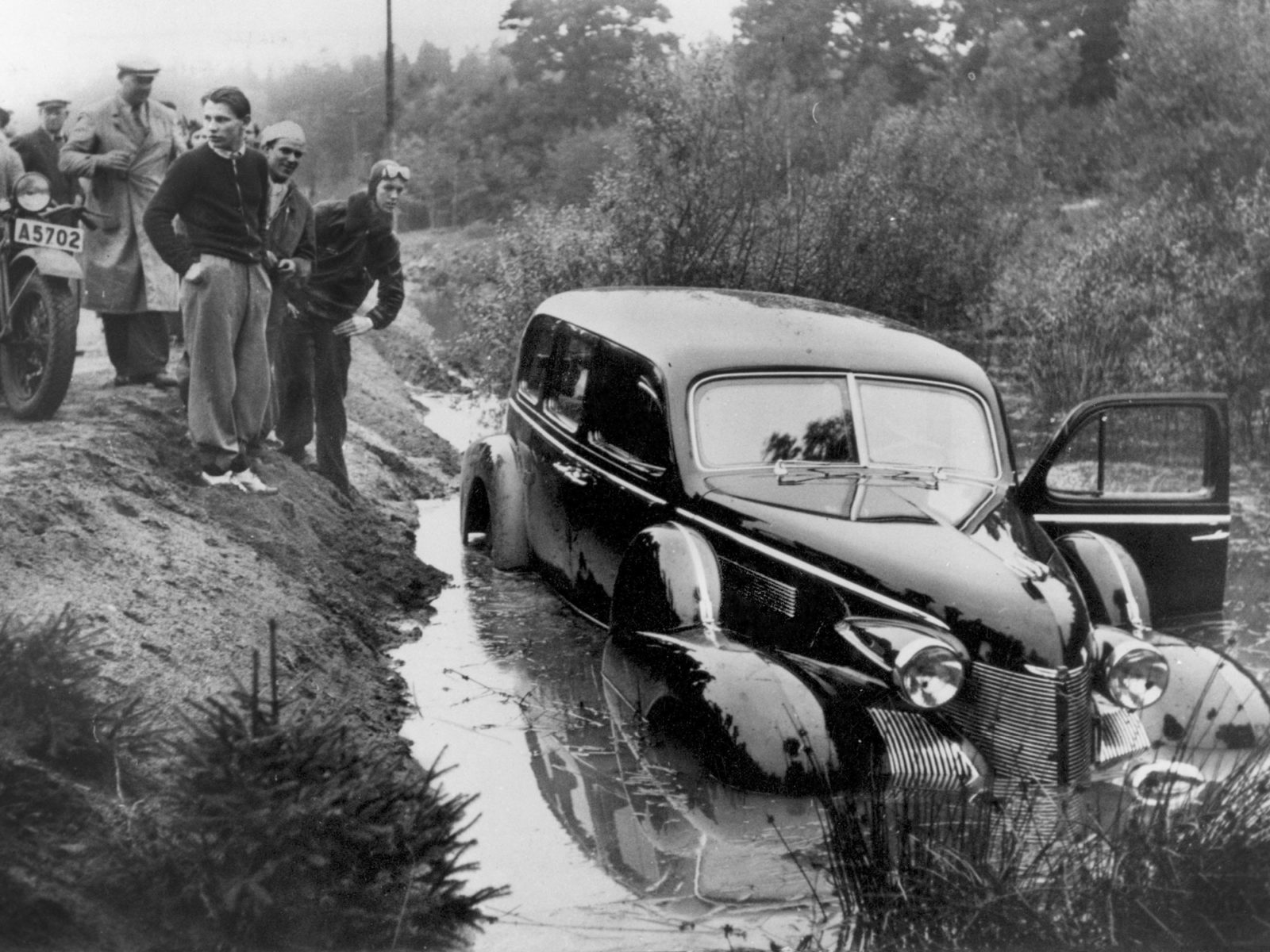 En 1940-tals-Cadillac står i vattenfyllt vägdike. Manliga åskådare i 1940-talskläder har samlats på vägen ovanför.