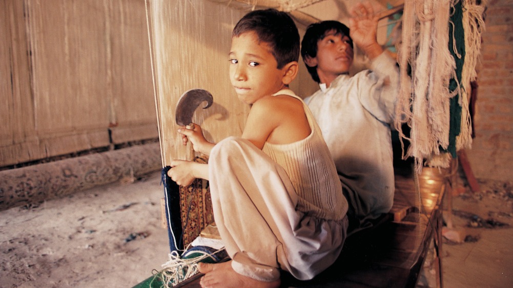 Boy squats at carpet loom, looking over his shoulder with a worried expression. Another working boy is seen in background.