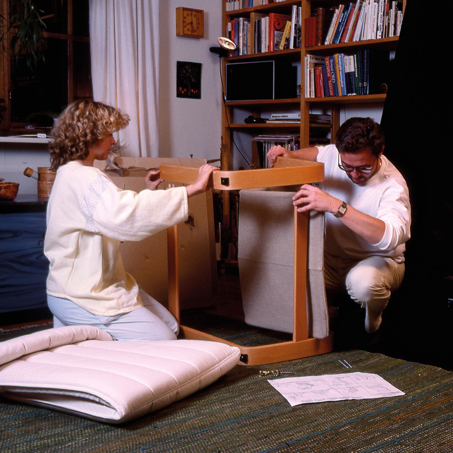 A blonde woman and dark-haired man sit on a green carpet, assembling a chair while man reads instructions.