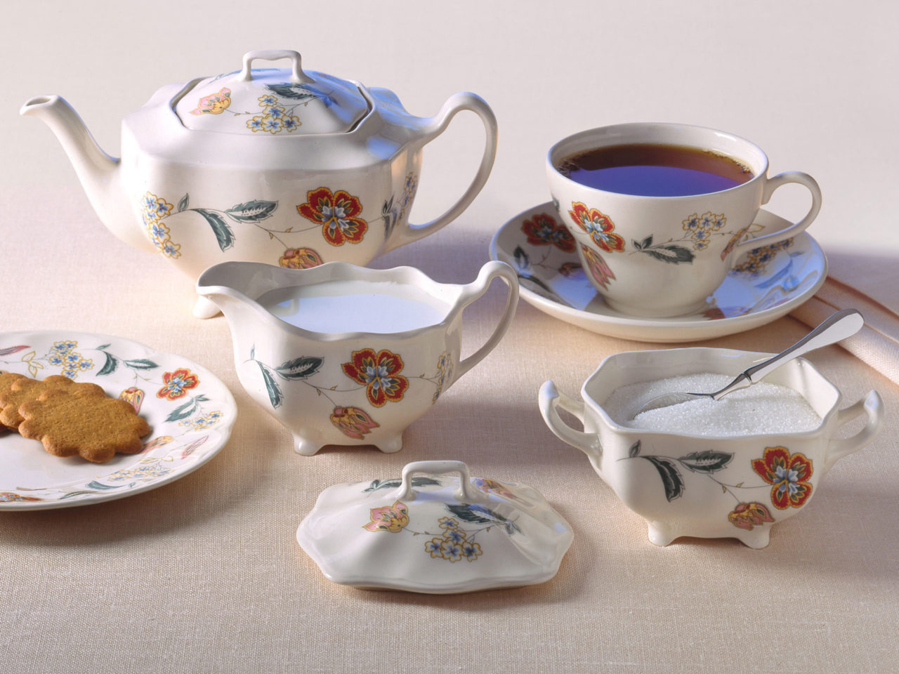 White crockery with multicoloured floral pattern, filled coffee cup, sugar bowl, creamer and plate with cookies.
