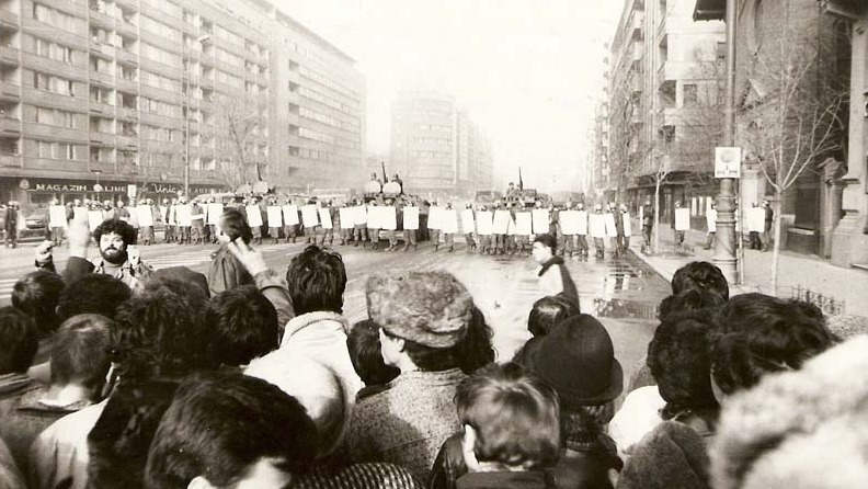 Manifestantes con ropa casual en una avenida urbana alzan sus puños frente a una fila cerrada de policías.