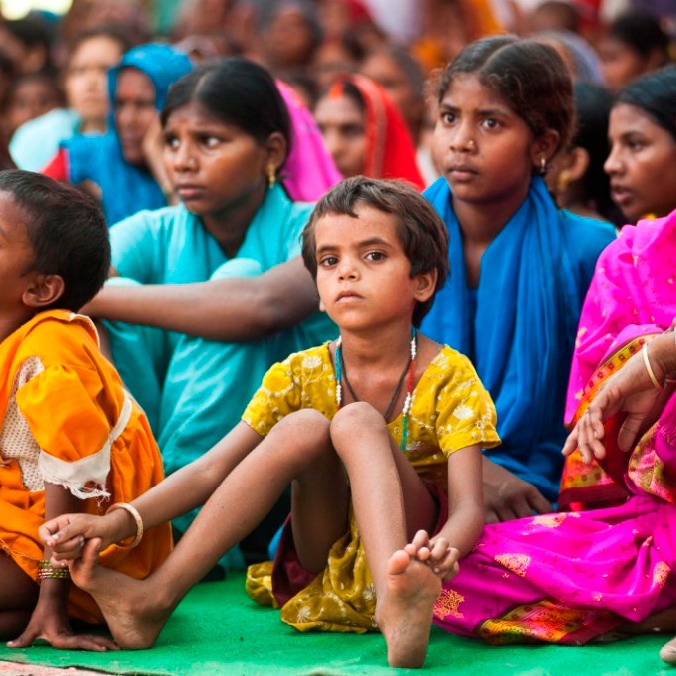 Folksamling med barn och kvinnor i färgstarka indiska kläder sitter tätt ihop på marken.