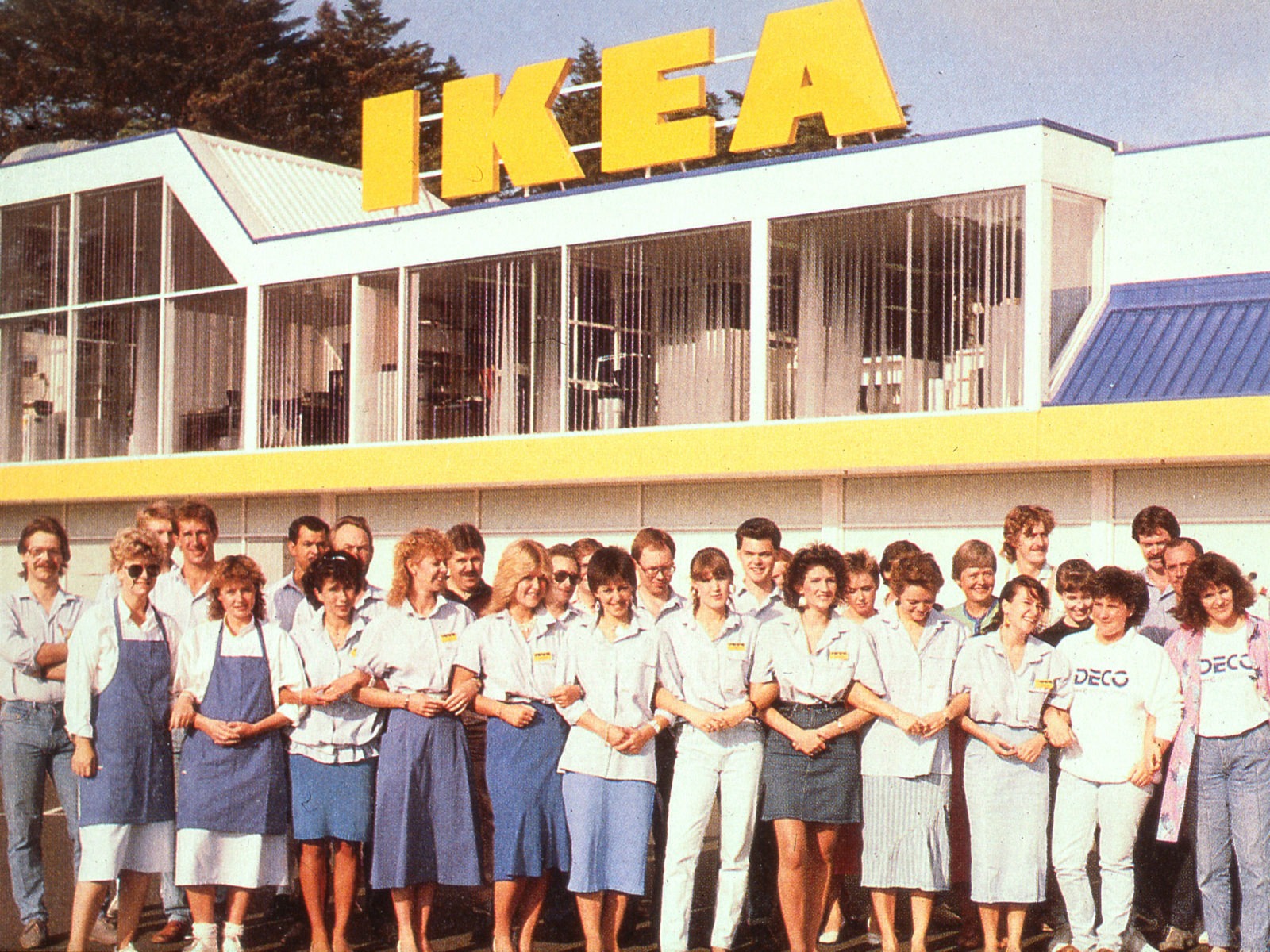 Group photo of IKEA staff in Germany wearing blue and white clothes in front of white building with yellow IKEA sign.