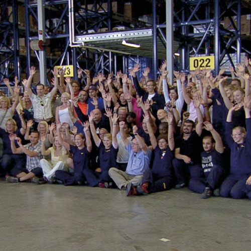 Stor grupp leende människor står eller sitter på golvet på IKEA tag-själv-lager.