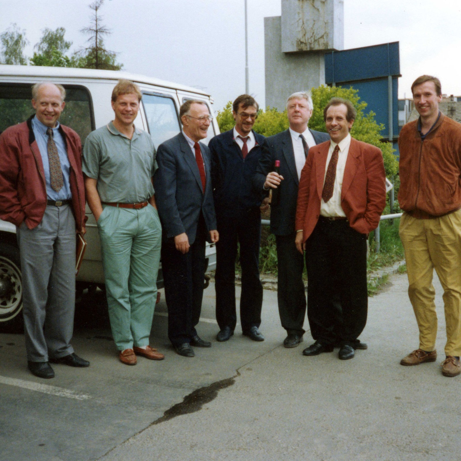 Sju män, uppklädda i 1980-talsstil, skrattar tillsammans på en parkeringsplats i ett fabriksområde.
