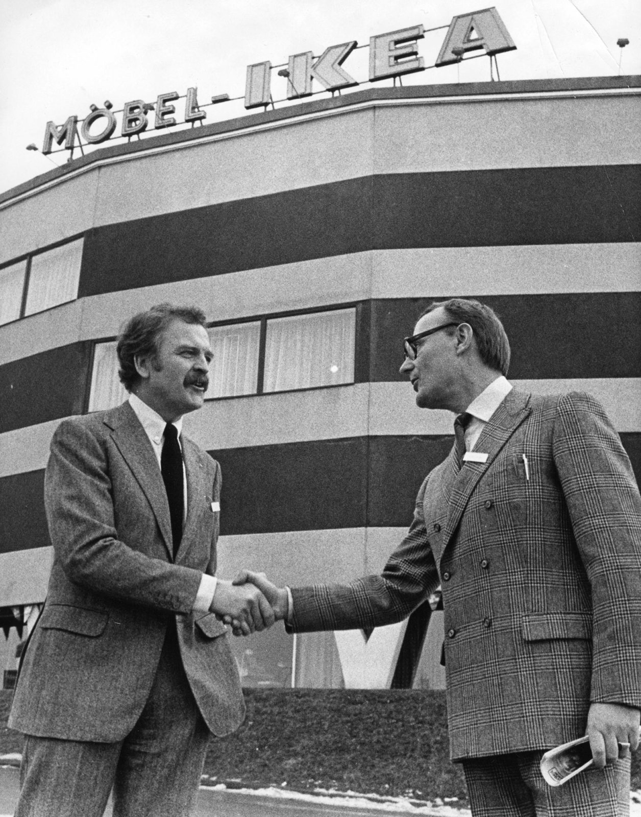 Hans Ax and Ingvar Kamprad in suits, shake hands in front of big building with sign FURNITURE IKEA in Swedish.