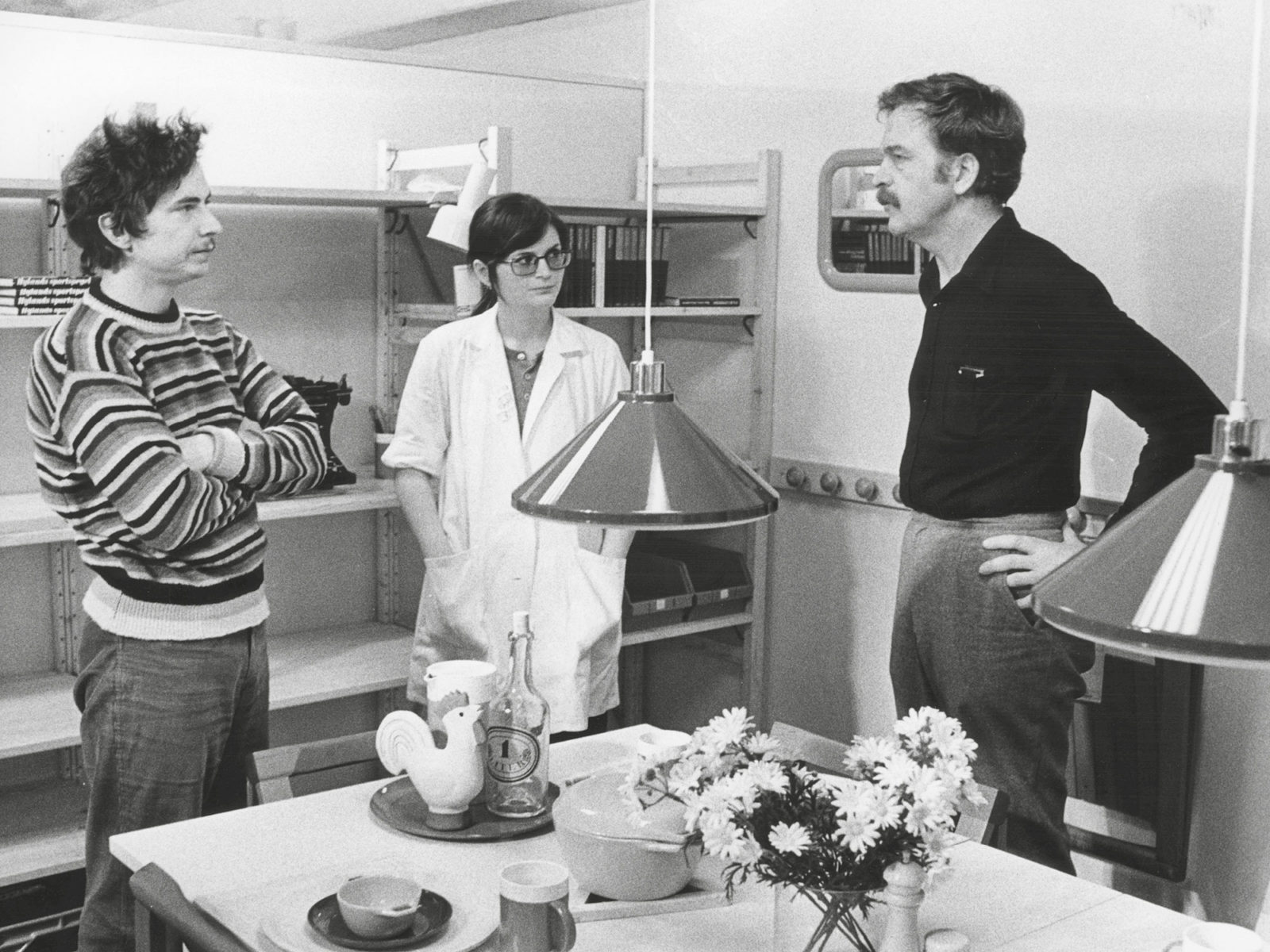 Tre personer står runt ett bord och pratar – Lennart Ekmark, Mary Ekmark och Hans Ax, ledigt klädda i 1970-talsstil.
