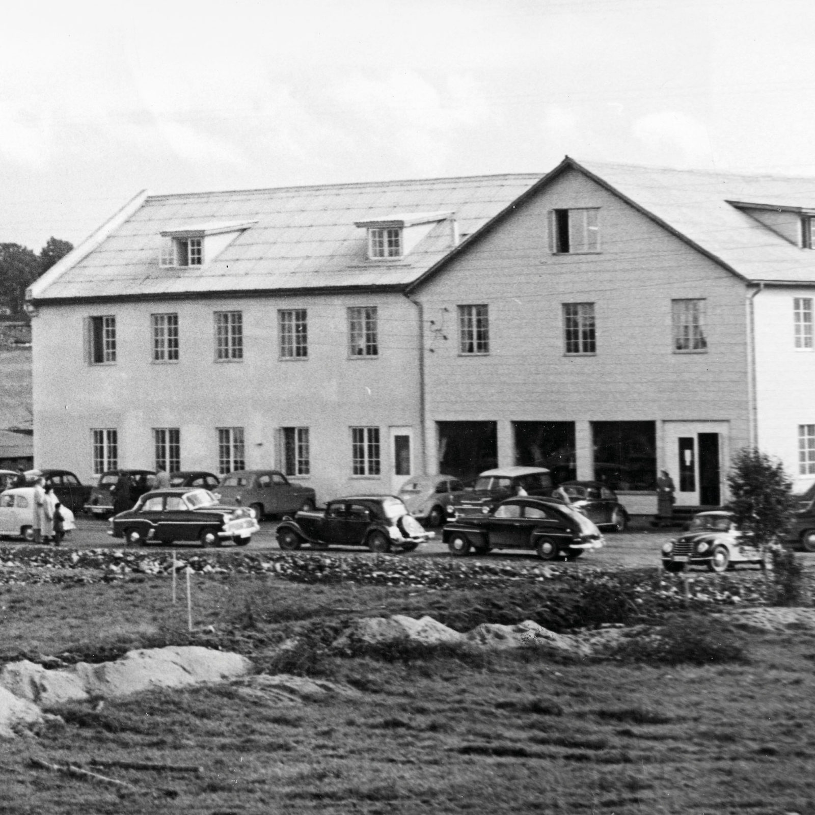 Black-and-white photo of big white building, 1940s and 1950s cars parked outside.