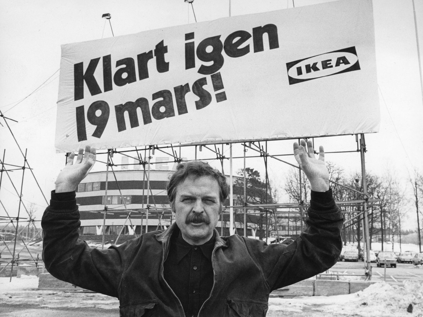 A moustached Hans Ax in suede jacket pretends to hold up huge sign on scaffolding with text: IKEA, Ready again 19 March.