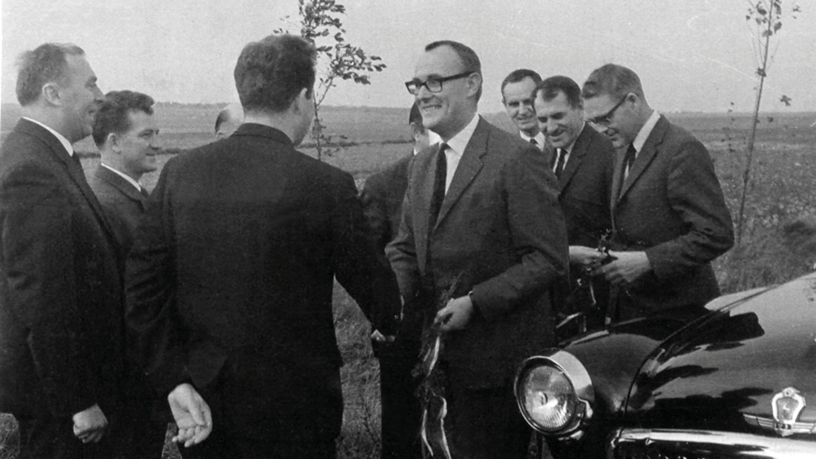 Grupp leende män i mörka kostymer står utomhus bredvid 1960-talsbil, en av männen skakar hand med Ingvar Kamprad.