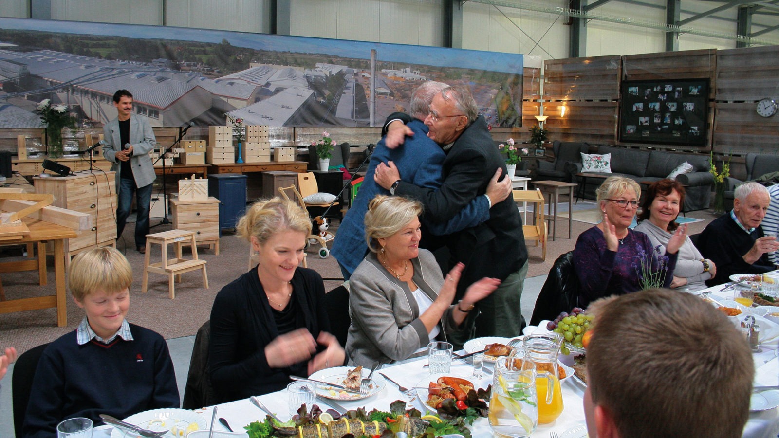 Group of happy people at festive table in factory showroom. Ingvar Kamprad stands and hugs Roman Prawda, both are in suits.