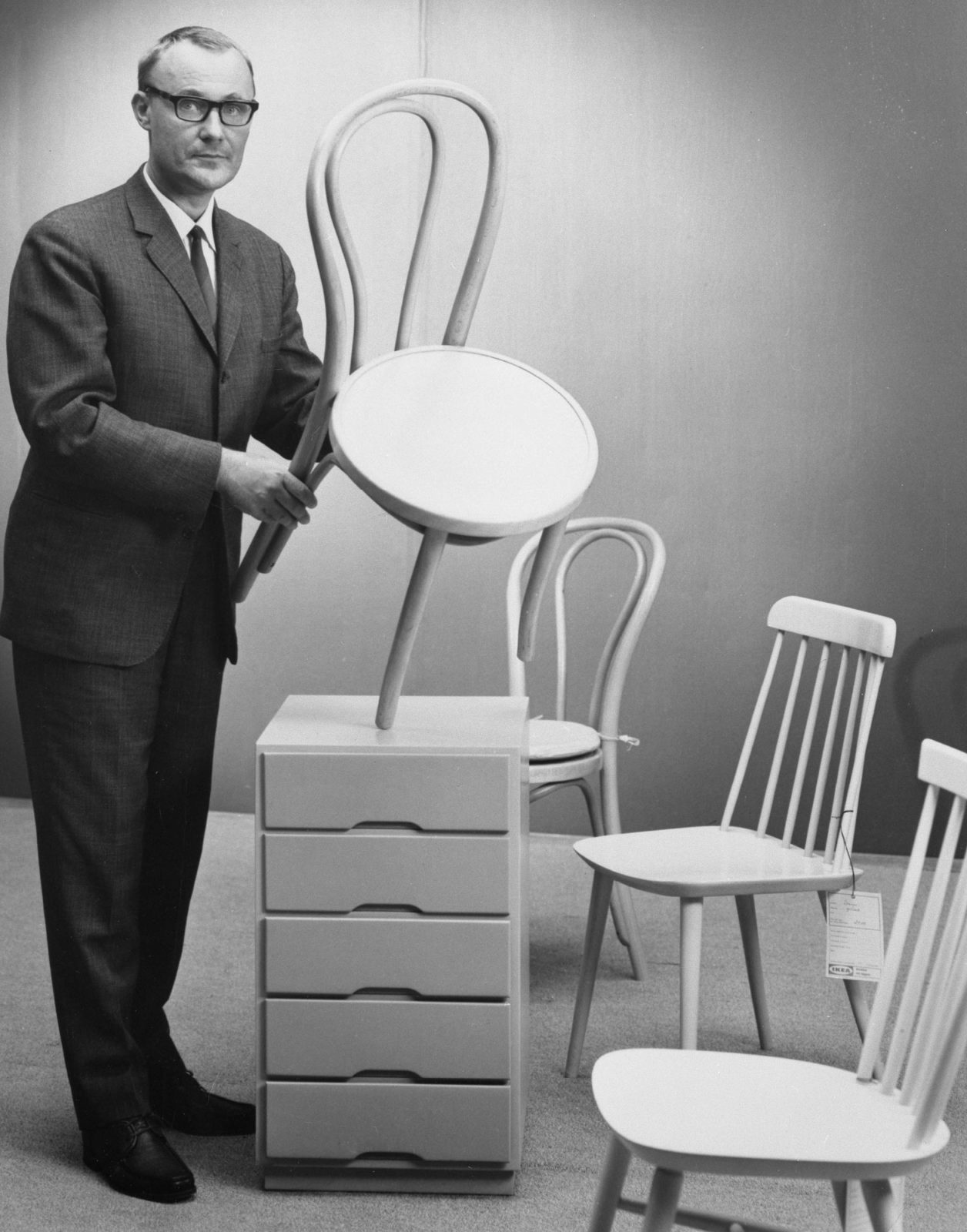 Ingvar Kamprad holding up white chair, next to 2 pin chairs and dresser, all white.