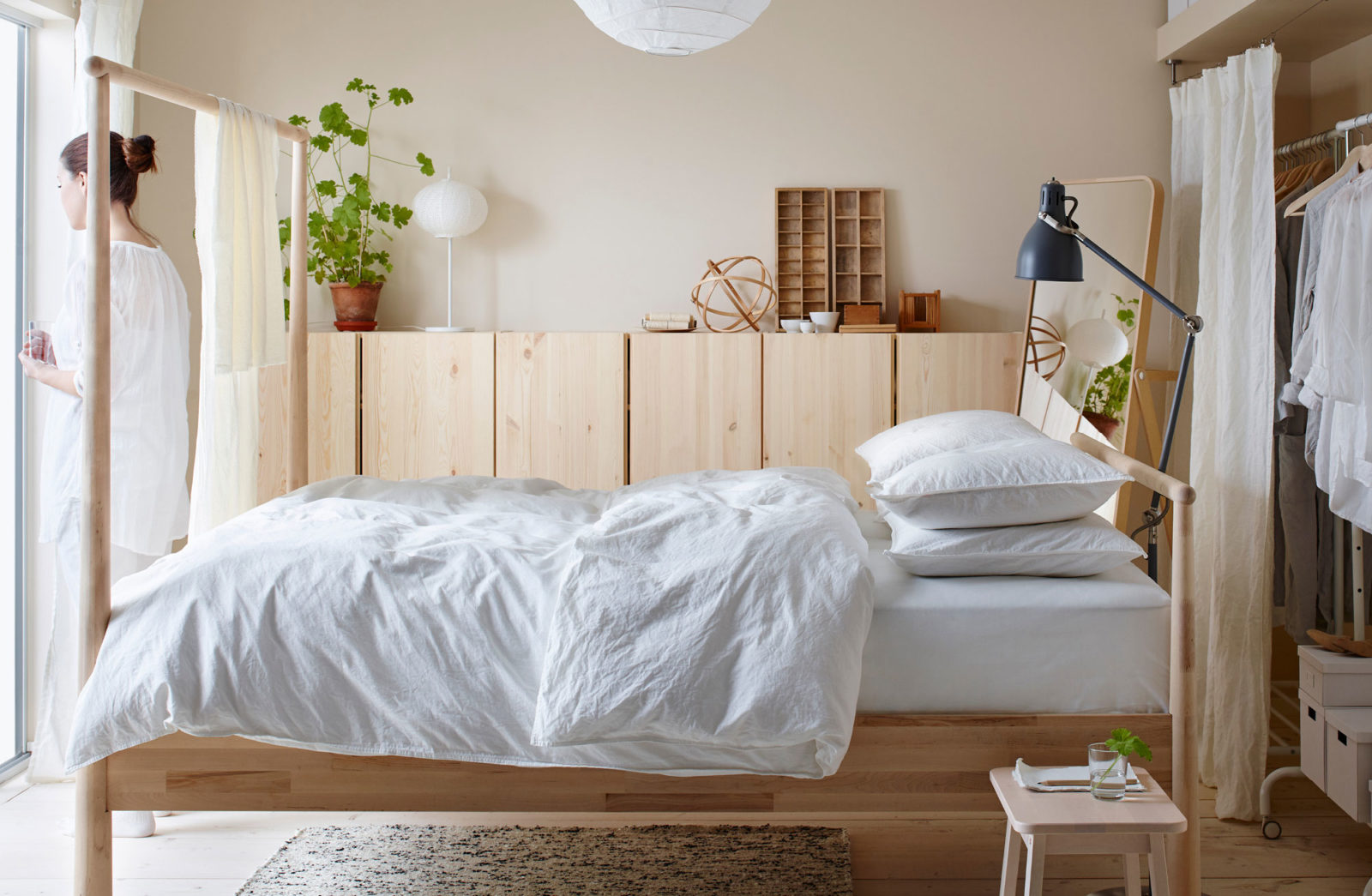 Scandinavian style bedroom with blond wood furnishings, white bed linen, a woman stands by bed with a glass of water.