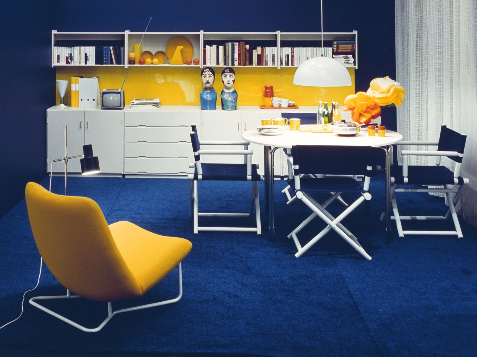 1970s style interior with blue carpet, white table, blue director's chairs, yellow armchair and white shelving system.