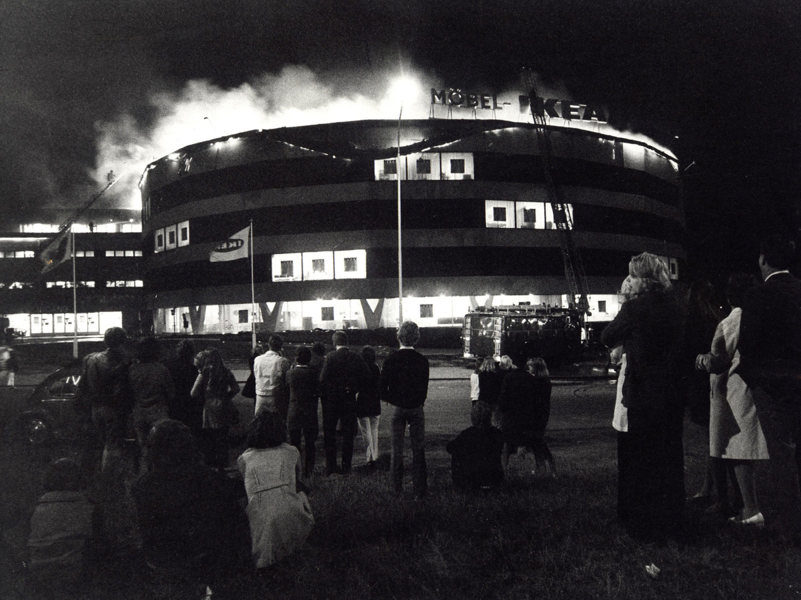 Åskådare har samlats nattetid framför enorm brinnande byggnad med skylten MÖBEL IKEA. En brandbil skymtar.