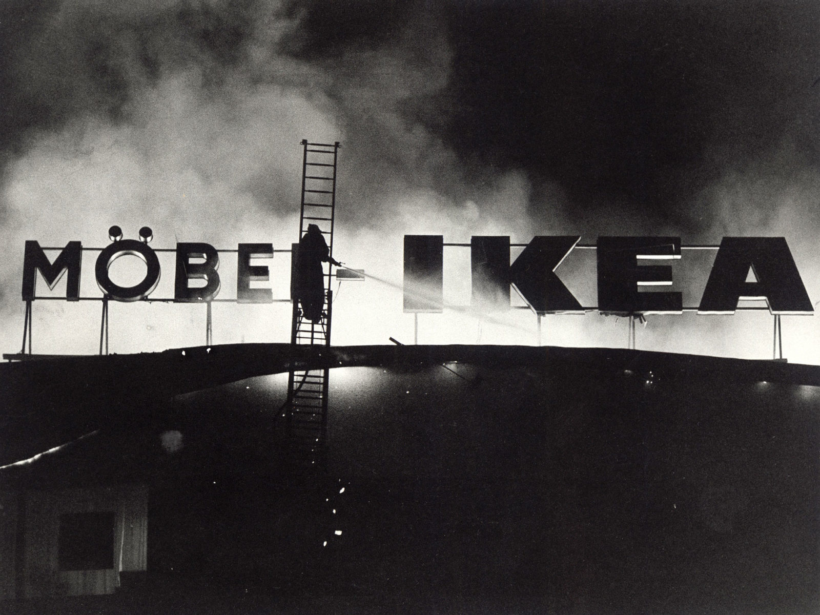 En brandman står på en brandbilsstege och sprutar vatten på ett neonskylt med texten MÖBLER IKEA, omgiven av rök.