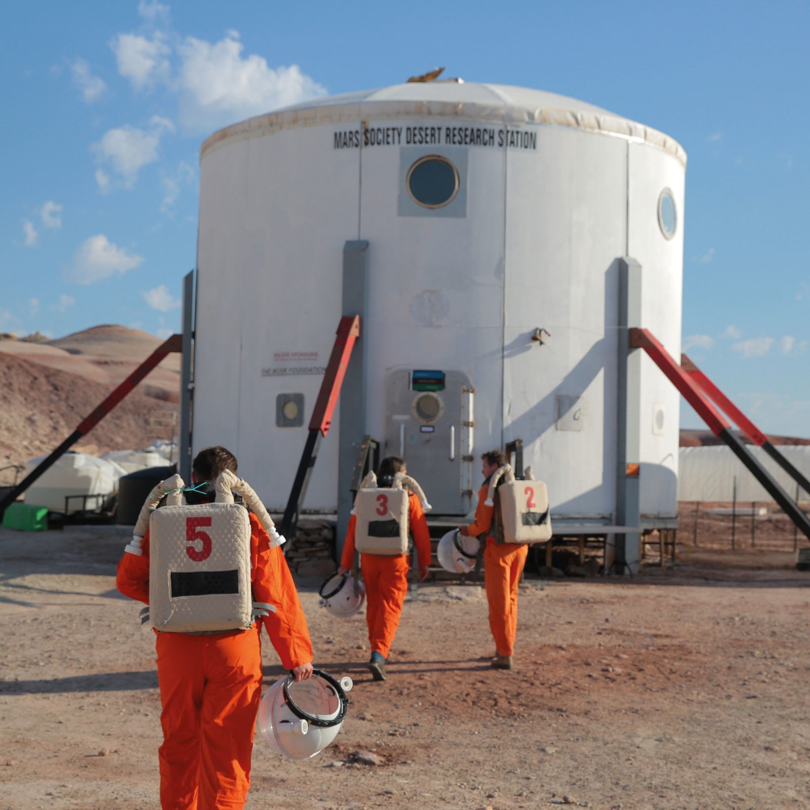 Three people carrying space helmets, dressed in orange jumpsuits, walk towards round grey metal building in the desert.