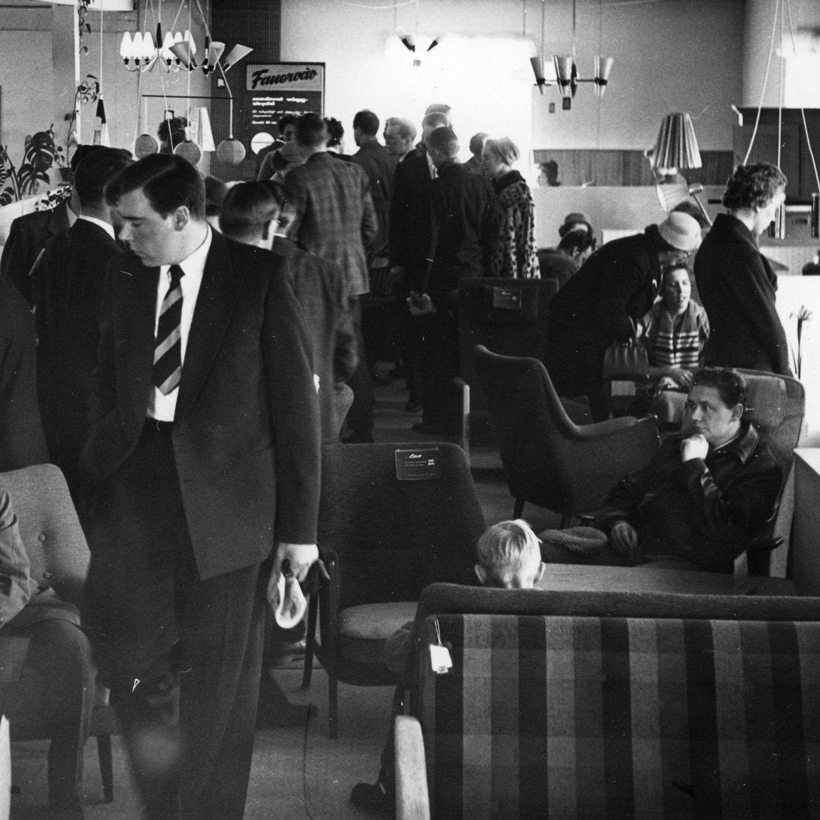 Svartvitt foto av möbelutställning där folk i 1950-talskläder trängs ochtittar på möbler och lampor.