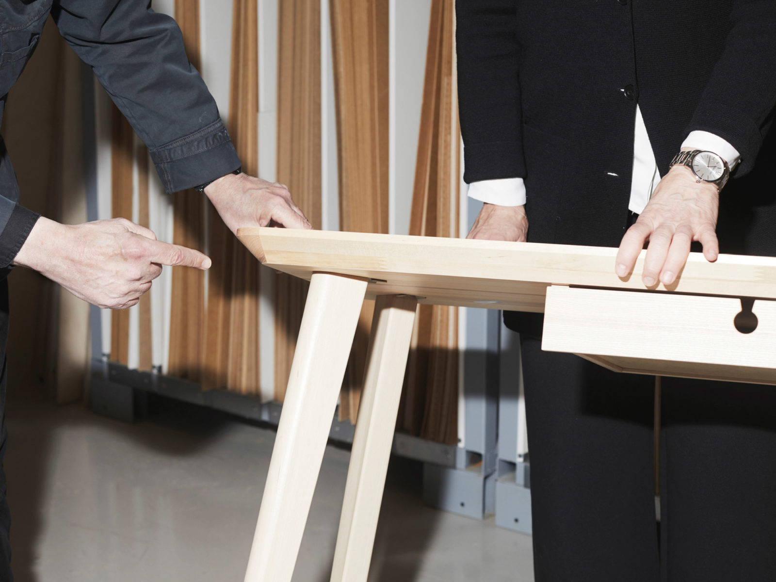 Close-up of two people holding a table, one pointing towards the area where a table leg is fastened on table top.