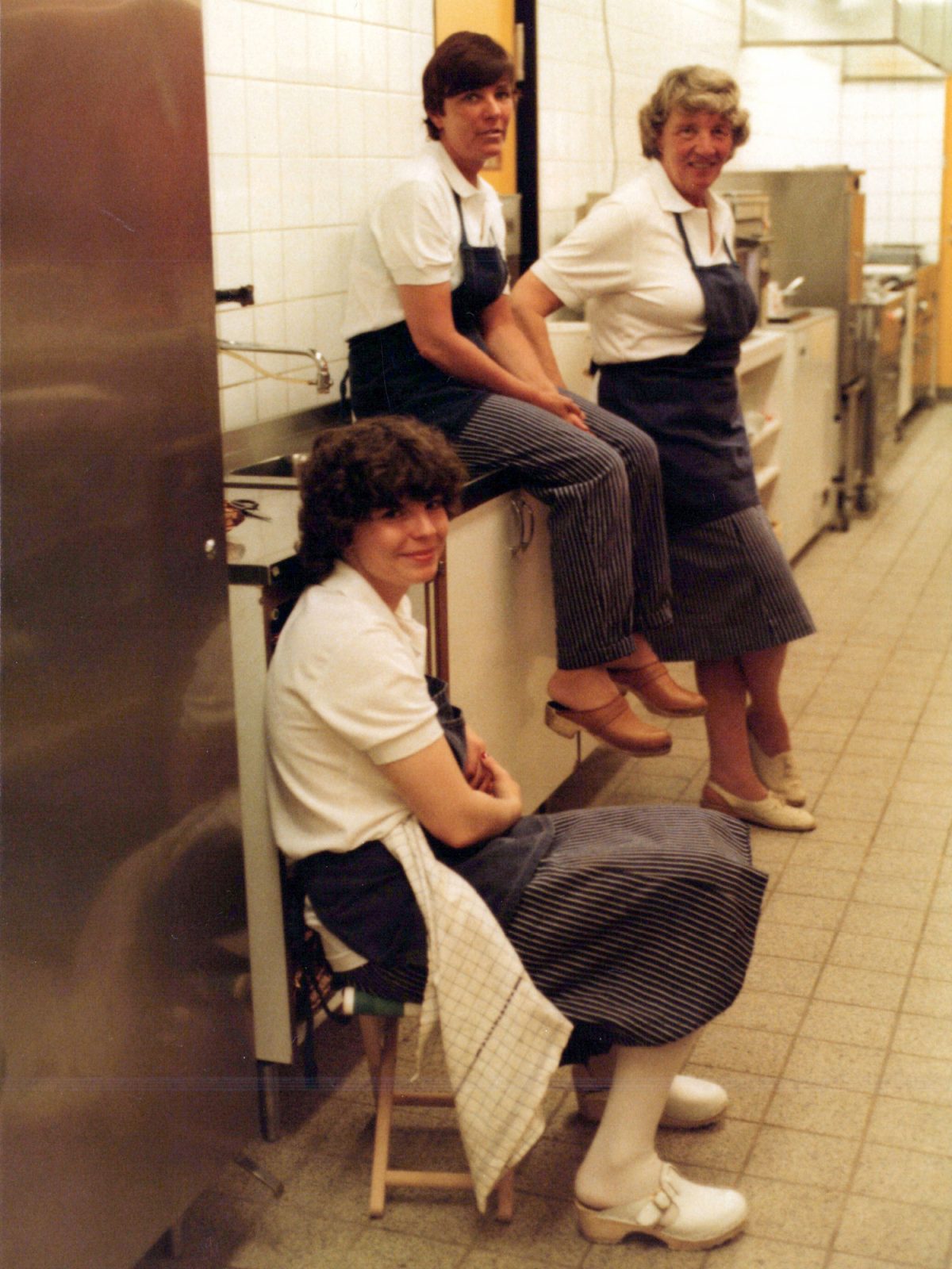 Three smiling women in industrial kitchen dressed in white with blue aprons.