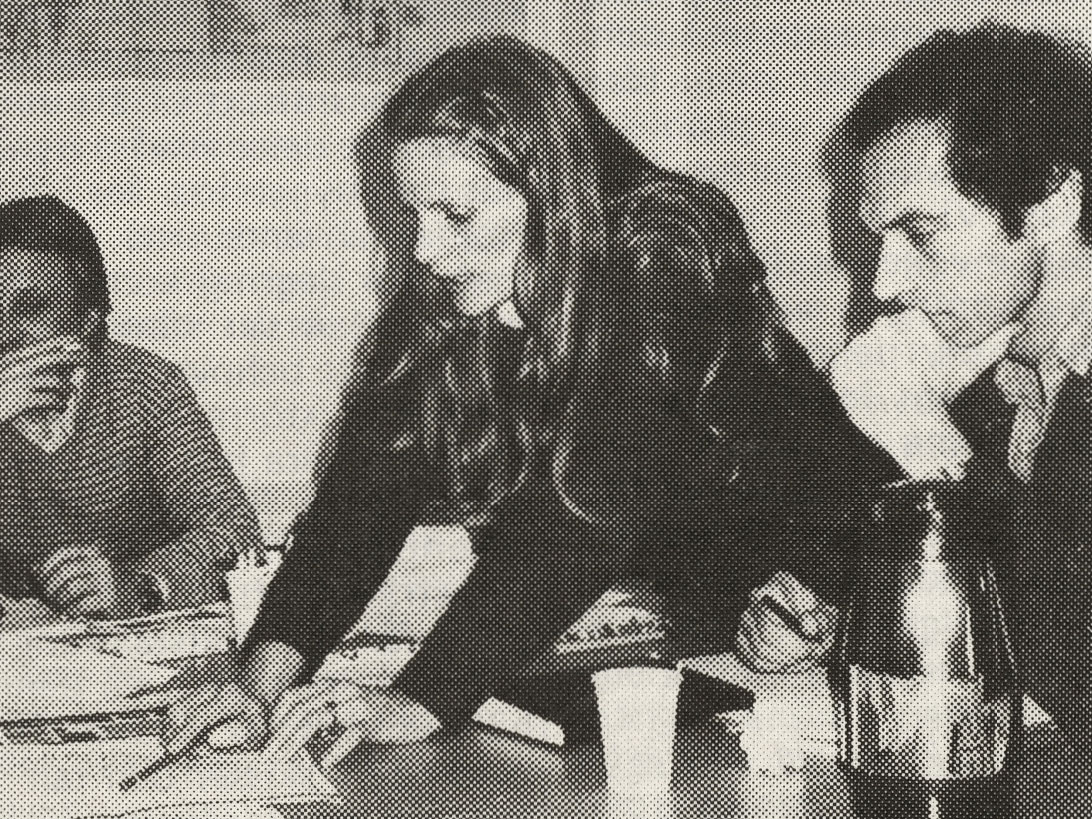 Tidningsbild av en kvinna och två män som arbetar tillsammans runt ett bord.