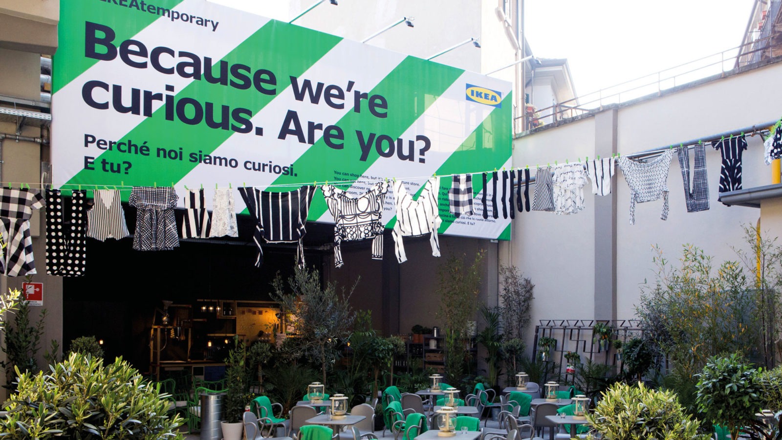 Garments on clothes line hanging over outdoor seating space, in front of big sign saying: Because we are curious. Are you?