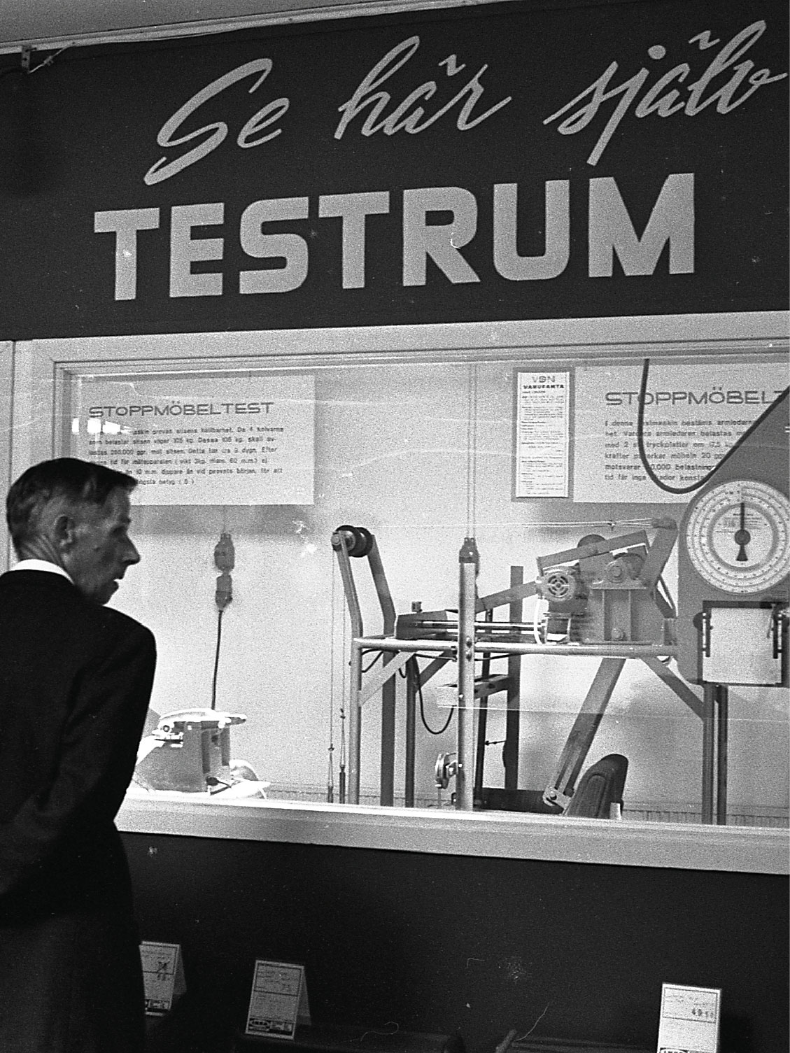 Unter einem Schild mit der Aufschrift „Überzeugen Sie sich selbst – Testraum“ schauen zwei Männer einer Möbeltestmaschine zu.