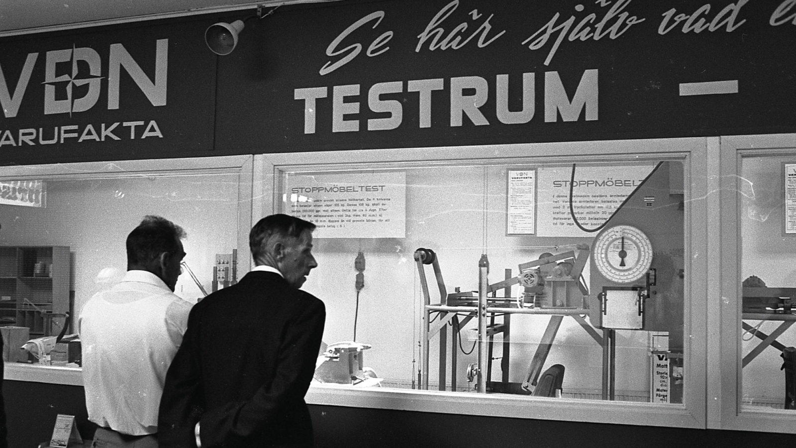 Unter einem Schild mit der Aufschrift „Überzeugen Sie sich selbst – Testraum“ schauen zwei Männer einer Möbeltestmaschine zu.