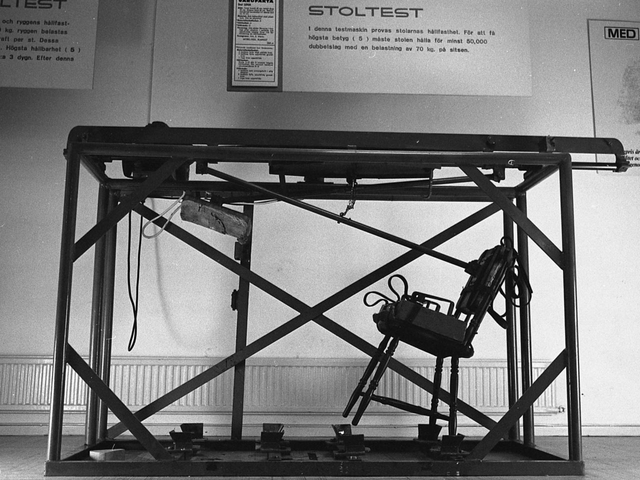 Large testing machine in which a chair is secured. Above is a sign with text 