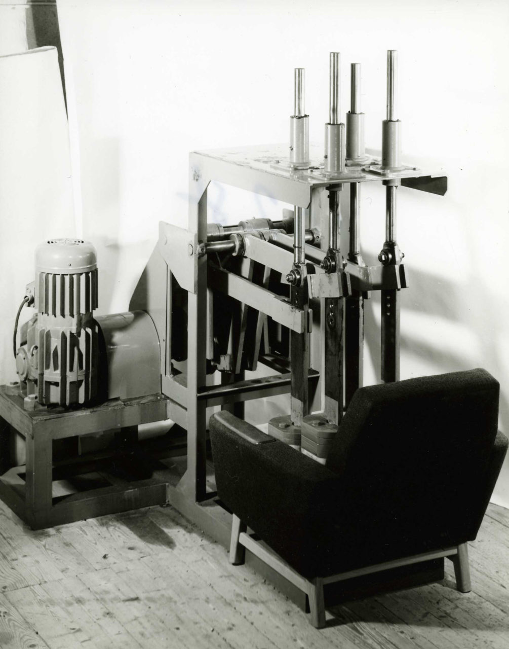 Große Testmaschine, in der ein schwarzer Sessel der 1960er dem Druck schwerer, pumpender Stahlkolben standhalten muss.