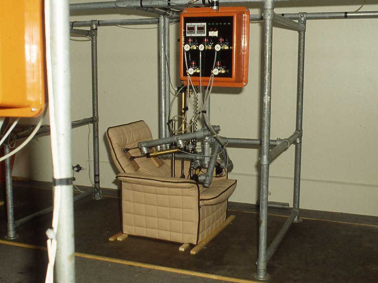 Une grande machine avec des tubes et des pistons en acier connectés à un tableau de contrôle frappant un fauteuil capitonné.