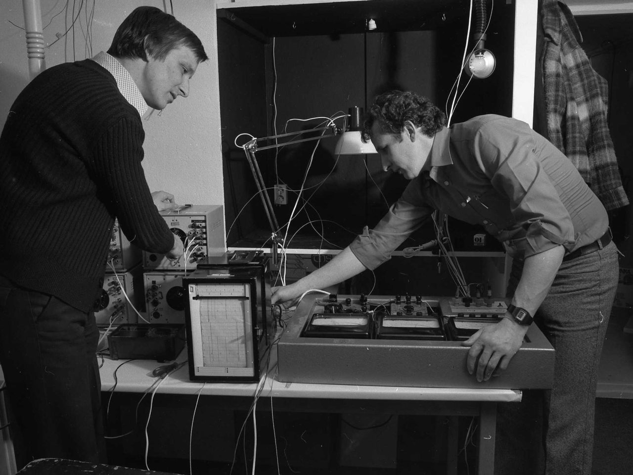 Two men dressed in 1970s style clothing check measuring instruments connected to a desk lamp.
