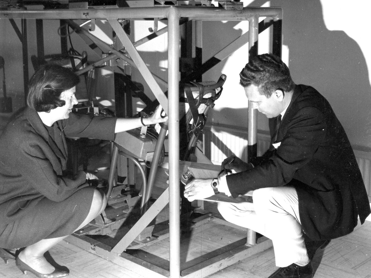 En kvinna och en man i 1960-talskläder, Karin Mobring och Nils Hasselstig, sitter på huk bredvid en maskin för möbeltestning.