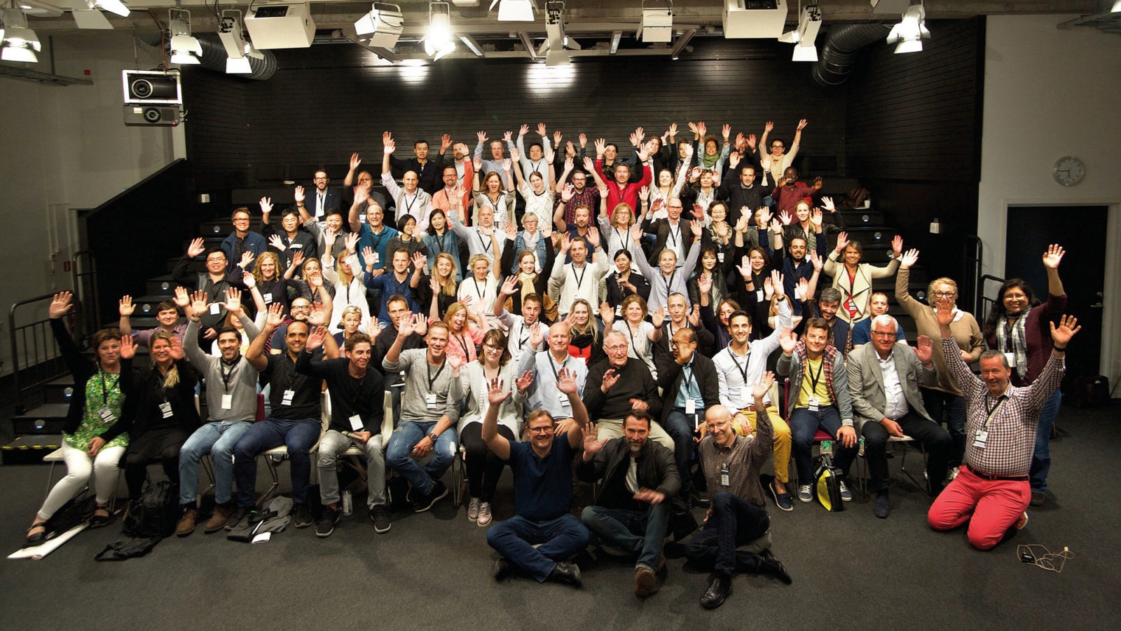 Stor grupp människor ler och vinkar i föreläsningssal. Ingvar Kamprad sitter i mitten på första raden.