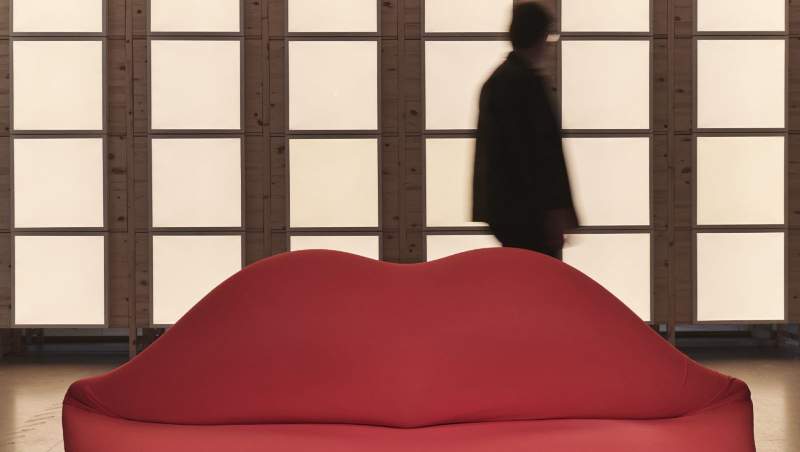 Ein rotes Bocca-Sofa in Form eines Mundes steht vor der Silhouette einer Person und einer Fensterwand.