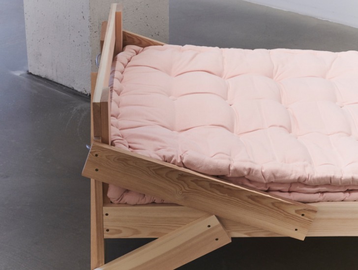 En ljusrosa madrass i en sängram gjord av träplankor som är slumpmässigt hopspikade, vilket ger möbeln ett rustikt och hemmasnickrat utseende.