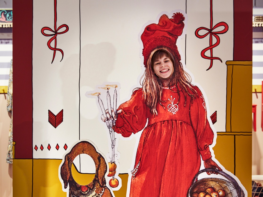 En leende kvinna tittar ut genom ett hål i målningen ”Brita som Idun” – en flicka i röd klänning tryckt på kartong med ett hål utklippt för ansiktet.