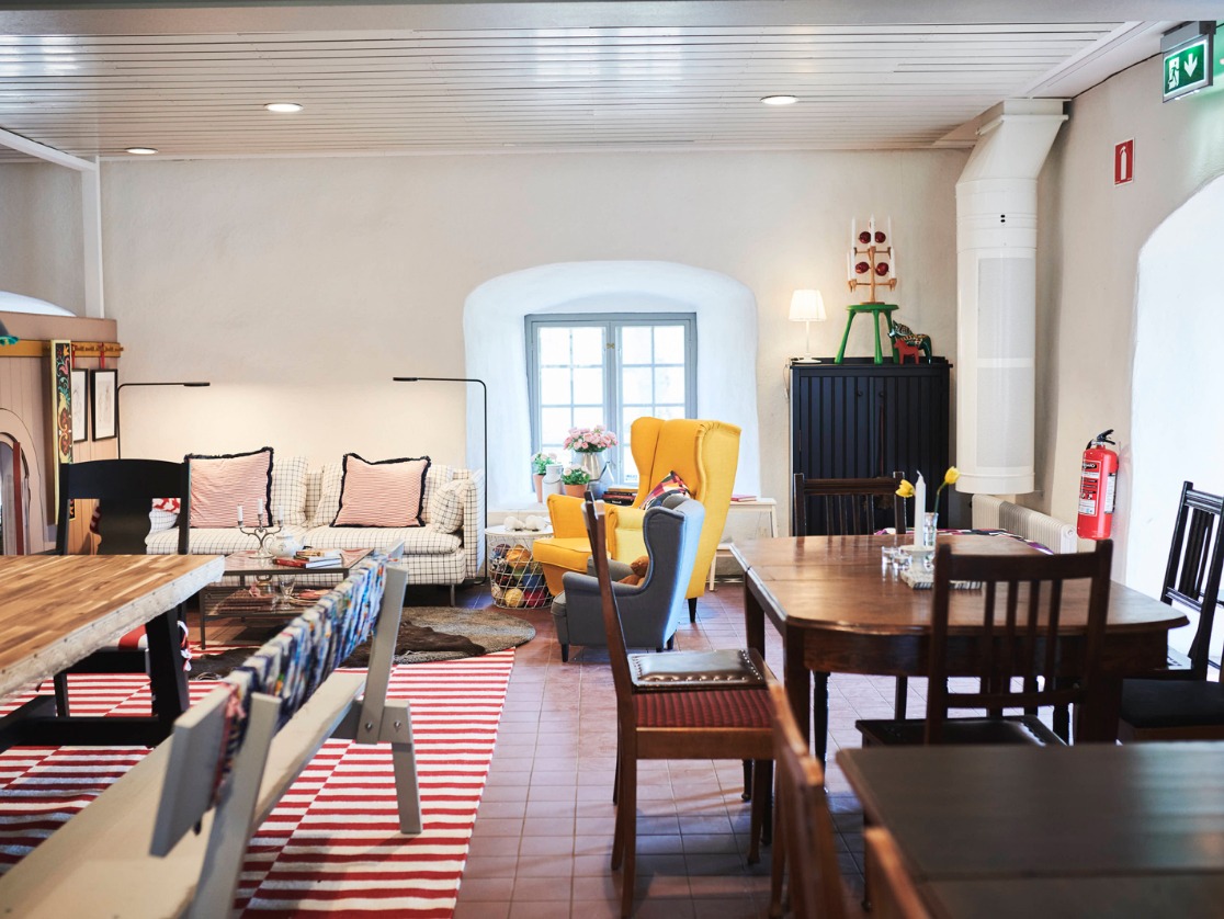 Ein großer Raum mit weißen Steinwänden, einem Schrank, zwei Ohrensesseln, Tischen, Stühlen und einer Bank im Stile Carl Larssons.