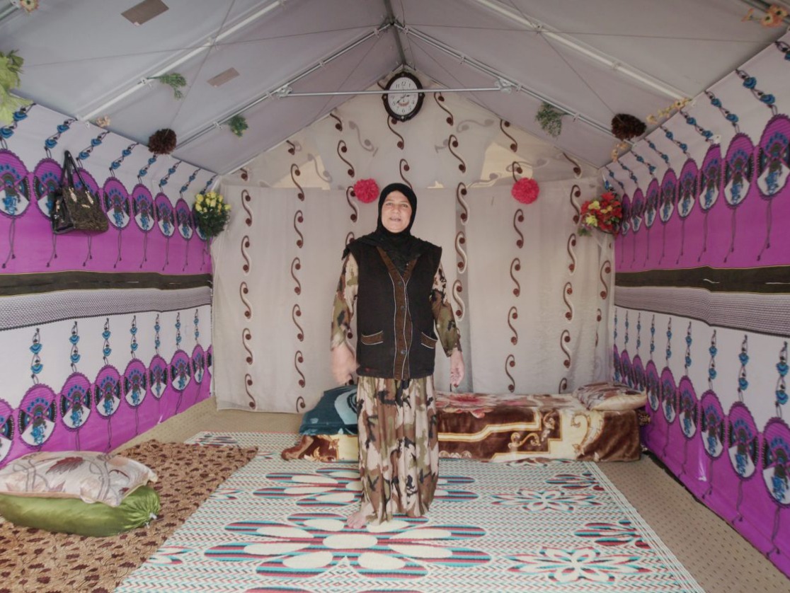 Eine Frau in ihrer Notunterkunft eines Geflüchtetenlagers, mit einem Bett, Kissen, einem Teppich und mit bunten Stoffen verzierten Wänden.