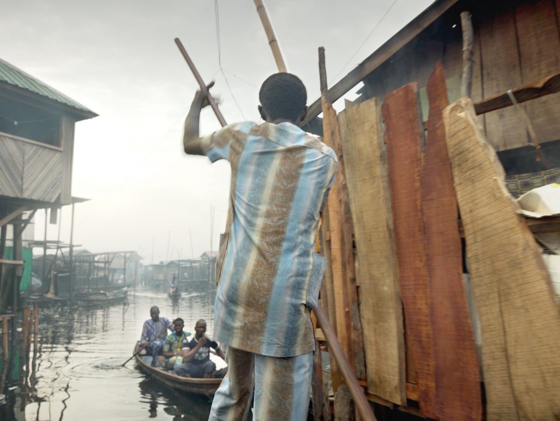 Häuser an einem Fluss in Nigeria und Menschen, die in kleinen Booten in verschiedene Richtungen rudern.